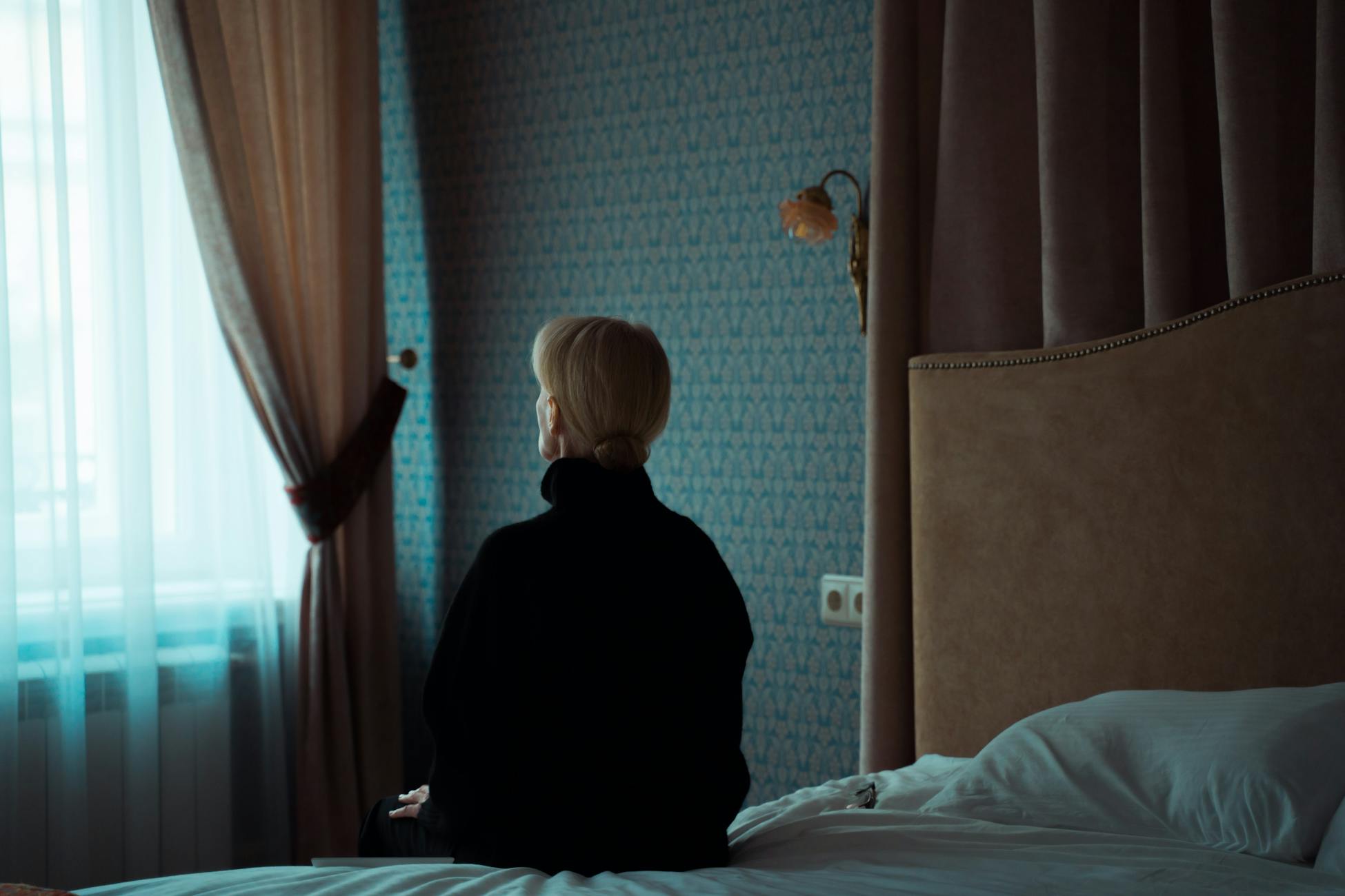 A woman in a dark room sits on a bed facing the window, reflecting solitude and contemplation.