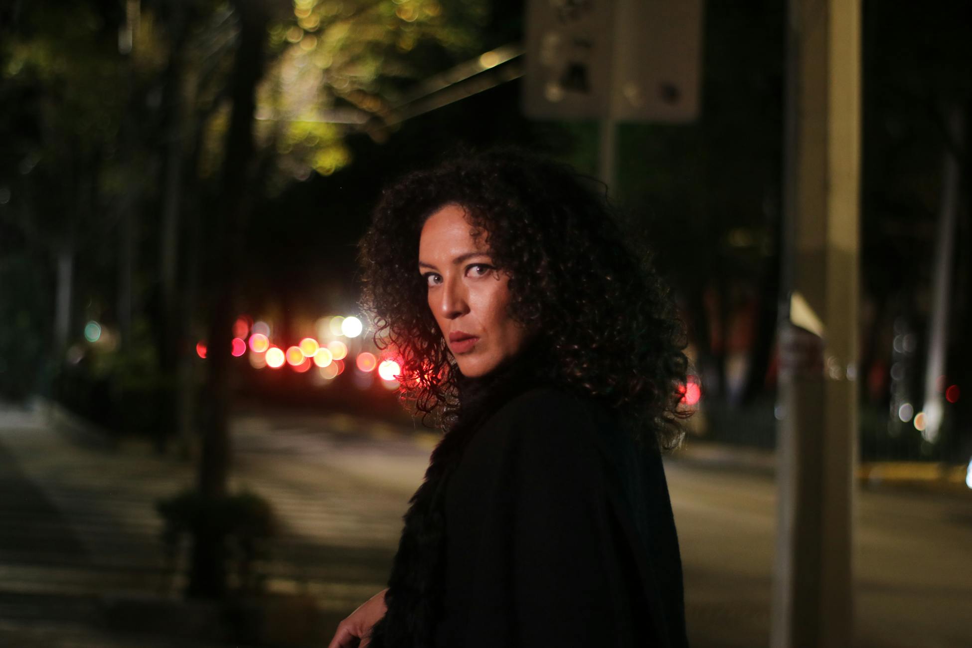 A woman with curly hair stands on a dimly lit street at night, looking back.