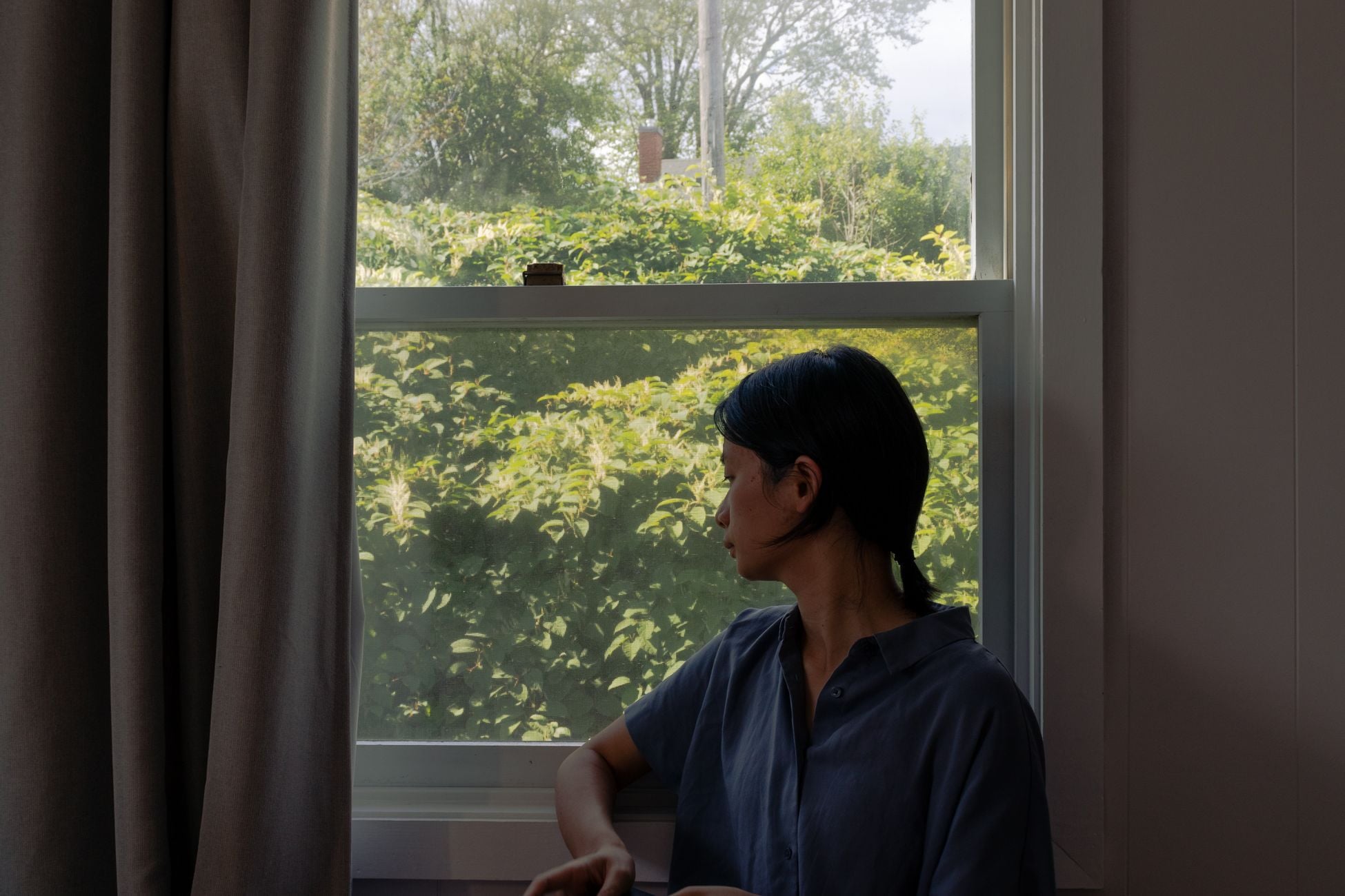 A young woman in casual attire looks outside through a window at lush green foliage.