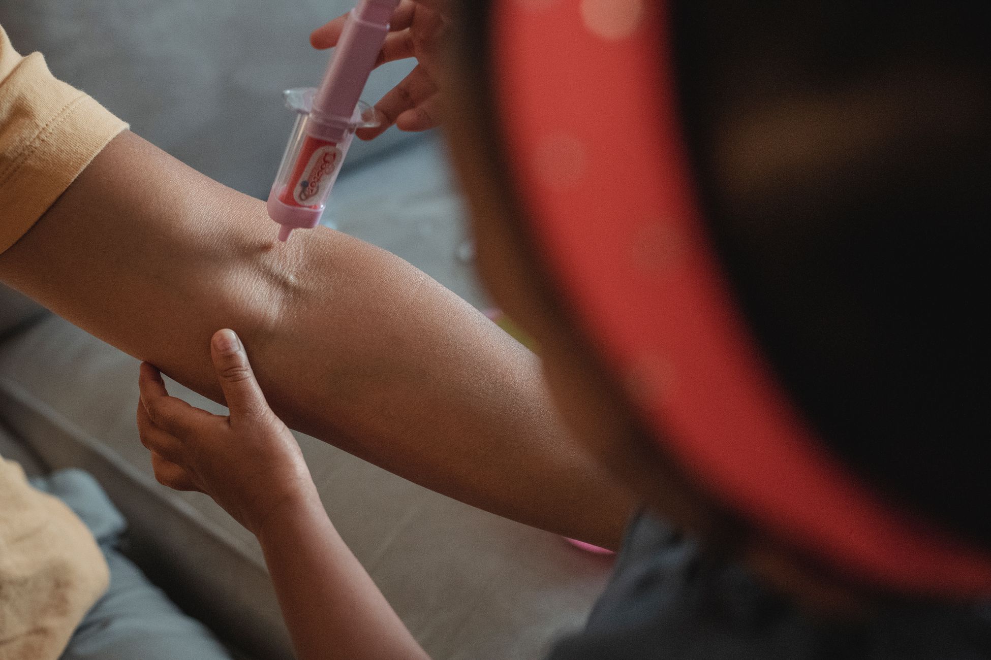From above blurred unrecognizable little ethnic girl with headband with toy syringe playing doctor with anonymous mother in apartment during weekend