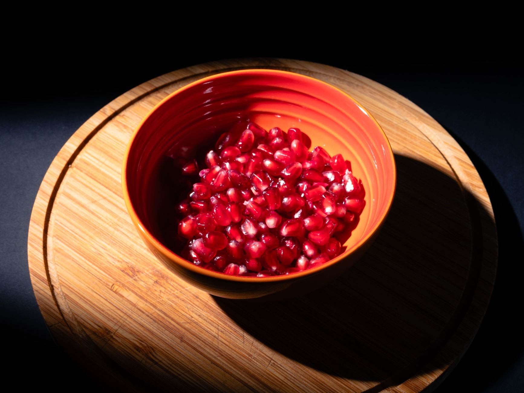 Fresh pomegranate seeds in a ceramic bowl on a wooden board, illuminated by dramatic lighting.