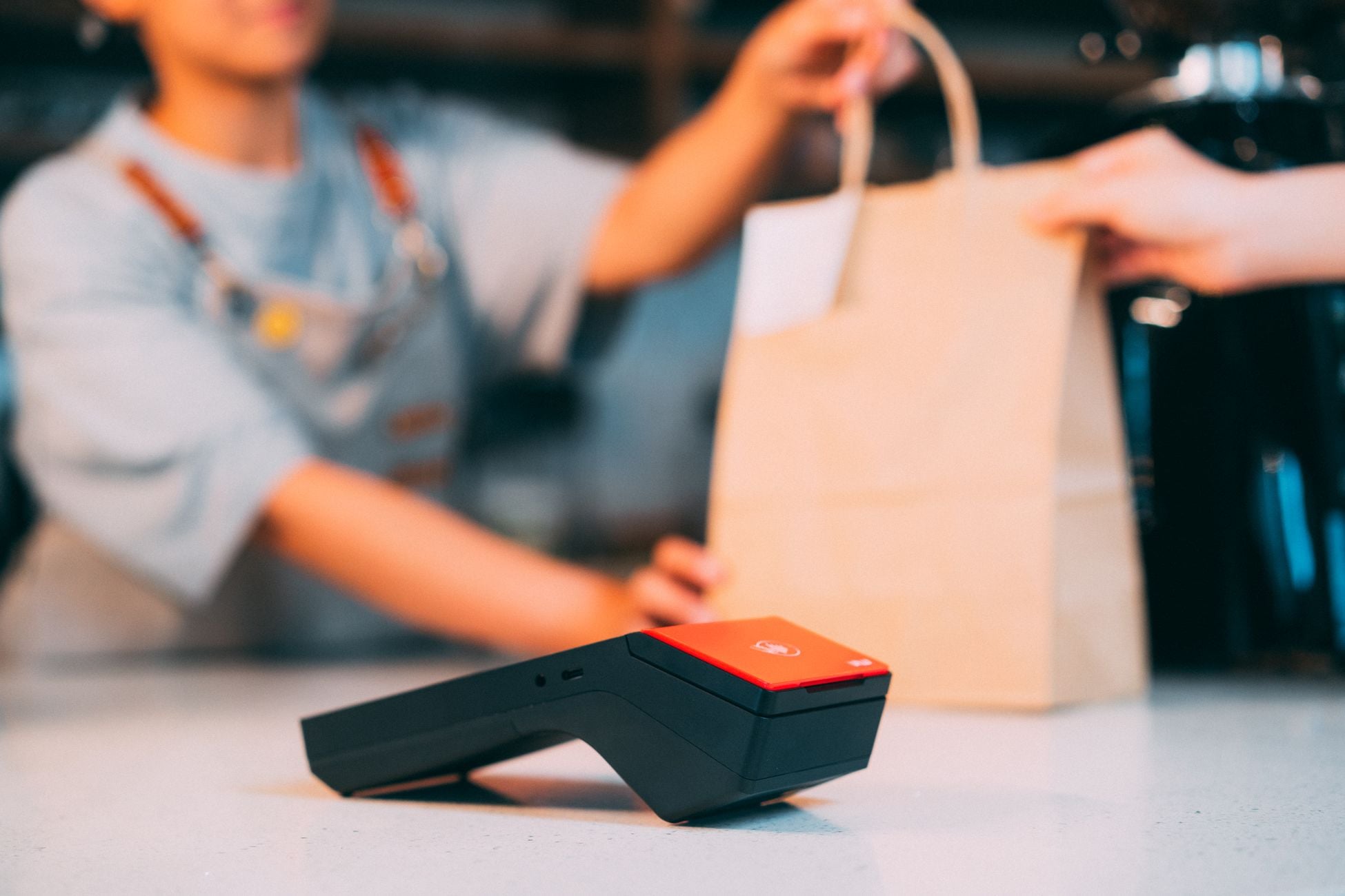 Cashier handing a takeaway bag to a customer with a contactless payment terminal in focus.